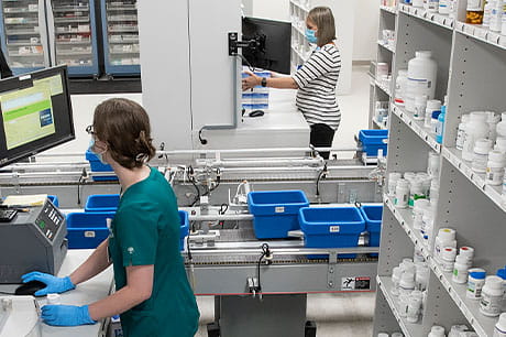 Pharmacy staff working with automated dispensing equipment and organizing prescription medications in a clinical pharmacy setting.